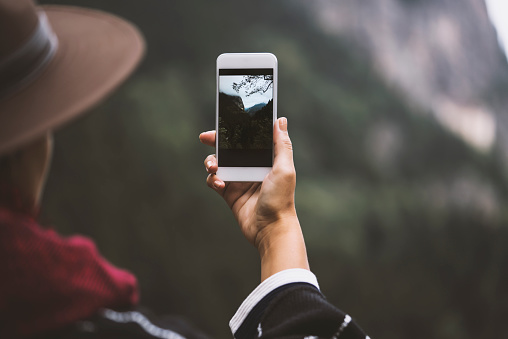 Young woman wearing a cape with a cowboy hat, taking photo with her mobile phone. Photographing the mountains and the forest over the hill under cloudy, rainy sky.