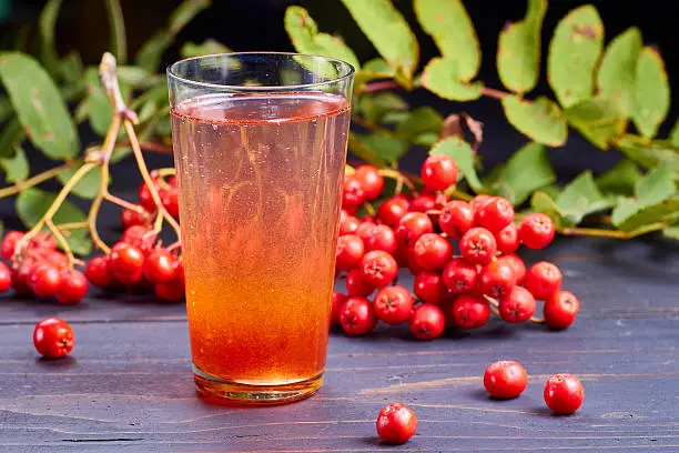 Glass of drink with syrup of rowan and fresh rowan berries on dark wooden table.