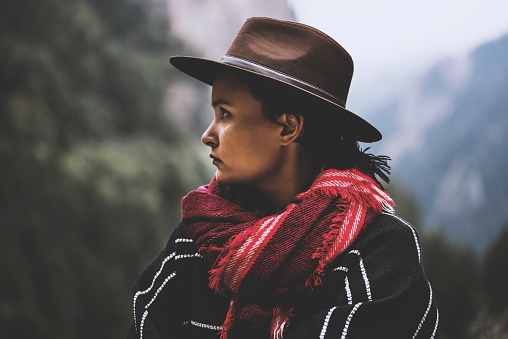 Portrait of young woman wearing a cape with a red scarf and a cowboy hat, looking away.  Forestry mountains at the background.