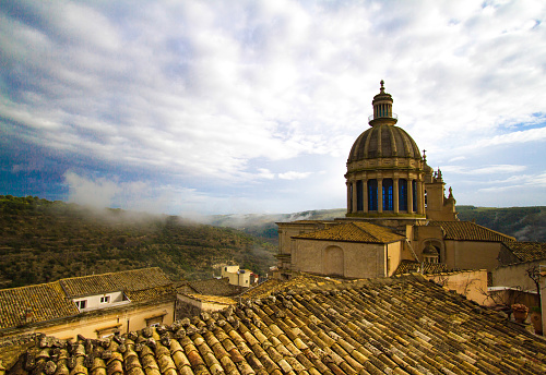 Ragusa Ibla, Sicily: Panorama with old tile rooftops and the baroque San Giorgio Cathedral under a cloudy-foggy-blue sky. Dating from the 1700s, the late baroque monument--designed by Rosario Gagliardi--is in the UNESCO World Heritage town of Ragusa Ibla, Val di Noto, Sicily, Italy.