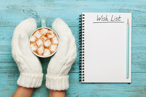Woman hands in mittens hold cup of hot cocoa or chocolate with marshmallow and notebook with wish list on turquoise vintage table from above, christmas planning concept. Flat lay style.