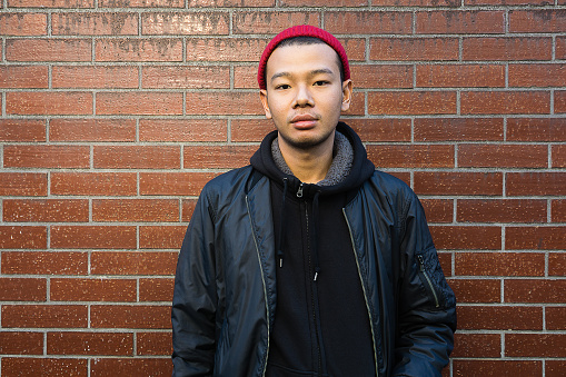 Handsome young asian man. Wearing black jacket and red beanie. looking at camera. Standing in front of brick wall