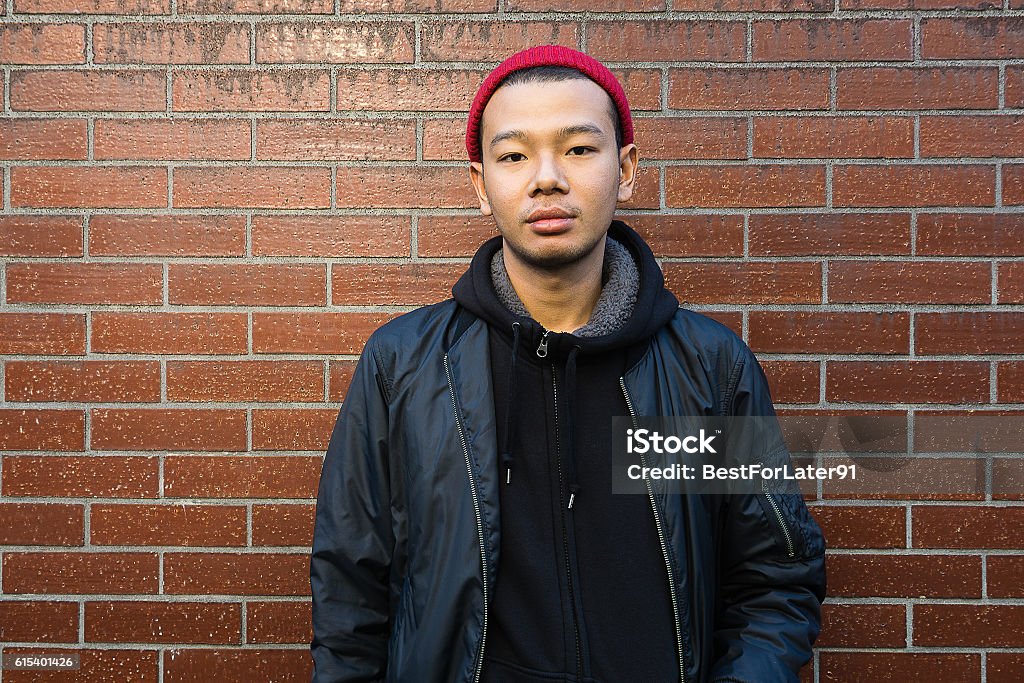 Handsome young asian man. Handsome young asian man. Wearing black jacket and red beanie. looking at camera. Standing in front of brick wall Teenager Stock Photo