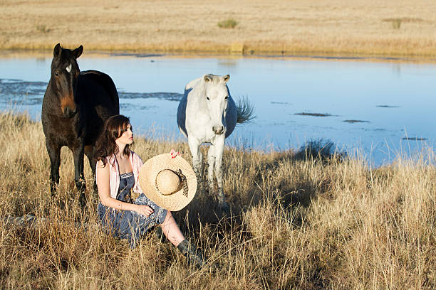 color portrait of a beautiful brunette woman in bohemian outfit with hat, seated in a field of grass with a stream, next to a dark and a white horse.