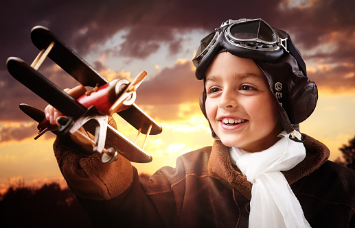 Boy wearing old-fashioned aviator hat, scarf and goggles holding a wooden biplane up in the air with sunset