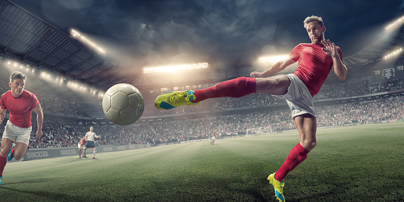 A professional male soccer player, dressed in a red and white soccer kit, in mid air about to kick a football in a volley. The footballer is playing with his teammates on an outdoor football pitch in a generic floodlit soccer stadium full of spectators at night. 