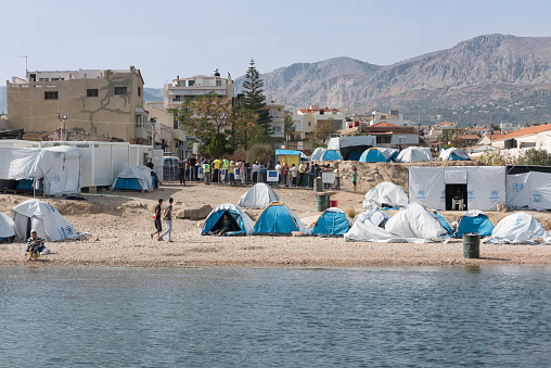 Chios, Greece - October 16, 2016: Refugees and volunteers (mainly from from the Norwegian NGO Drop in the Ocean) outside the Souda refugee camp in Chios Town on Chios Island in Greece. The main (official) camp area is overcrowded so refugees have started to place small tents on the beach outside (the camp area). In the background volunteers are serving lunch to refugees standing in line. At the moment 1,500 refugees are fed three meals each day.