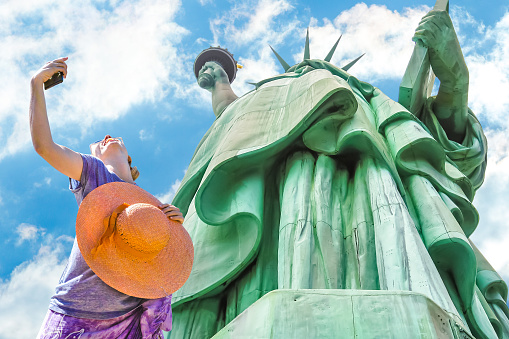 A smiling and fashionable woman with a orange wide-brimmed hat takes a selfie. Statue of Liberty and blue sky in the background. Liberty Island, New York City, United States