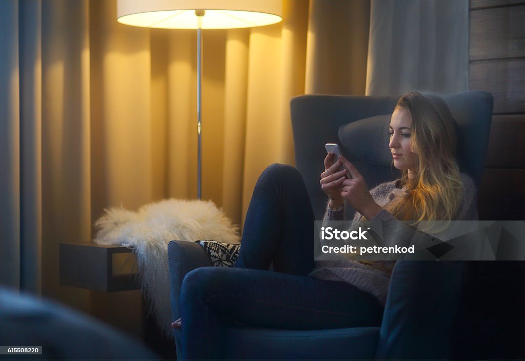 Woman typing text message on smart phone Woman lying on armchair while typing a text message on her cell phone Electric Lamp Stock Photo