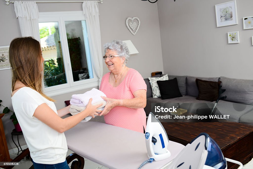 cheerful young girl ironing and helping elderly woman at home cheerful young girl ironing and helping with household chores an elderly woman at home Laundry Stock Photo