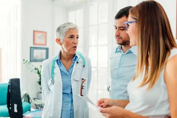 Pregnant woman and her female doctor in a consultation. Woman standing and holding ultrasound image and listening her doctor. Young woman husband is beside her.