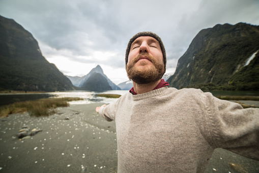 Young man stands arms outstretched in front of the Mitre peak in Milford sound, New Zealand. Concept of freedom and success.
