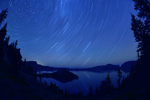 Wide angle, night view of the Crater Lake, a collapsed, volcanic caldera, Oregon, USA. Stars appear rotating around the North Star during long time (45 minutes) night sky exposure creating the star trails effect. Polaris is a bright dot in the upper-left corner.