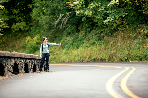 Ethnic adult female hitch hiker waiting for a ride on the side of a wooded highway