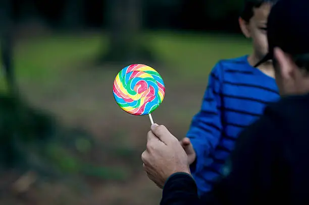 Strange adult male offering a young boy a lollipop as he tries to lure him from the park