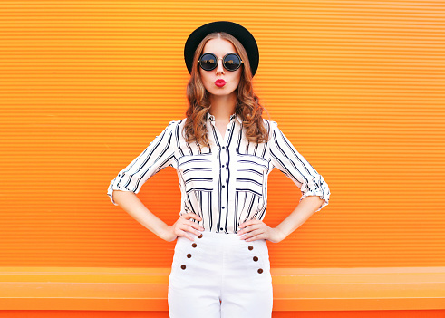 Fashion pretty glamour woman wearing a black hat sunglasses white pants over colorful orange background