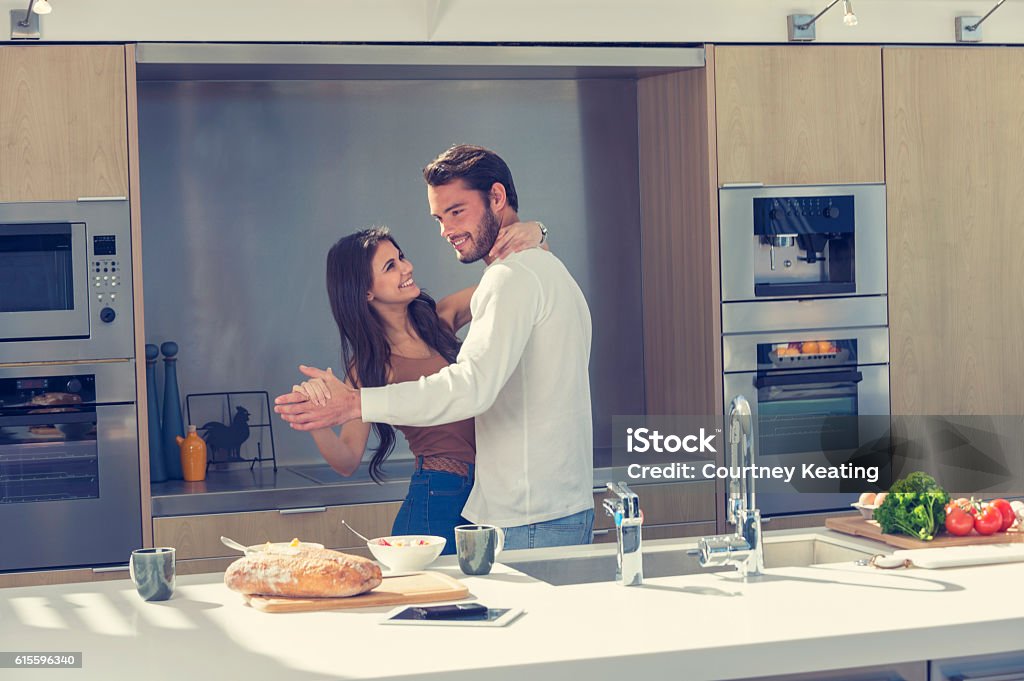 Couple playfully dancing in the kitchen. Couple playfully dancing in the kitchen. There is some food on the kitchen counter including bread and vegetables. There is also a digital tablet and mobile phone. Couple - Relationship Stock Photo