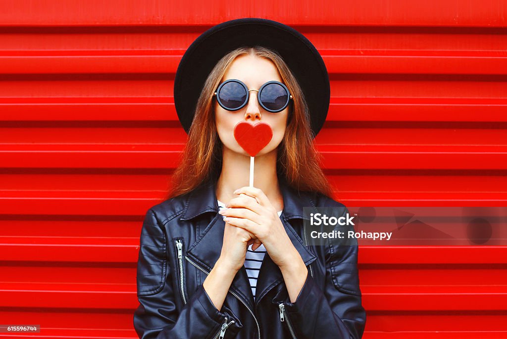 Fashion pretty young woman with red lollipop heart wearing black Fashion pretty young woman with red lollipop heart wearing black hat leather jacket over urban background Tasting Stock Photo