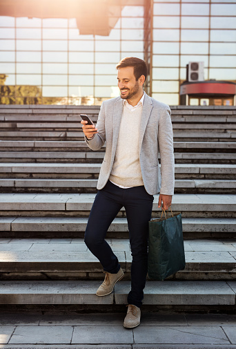 Young smiling man walking downstairs and reading text message on mobile phone while carrying shopping bag.