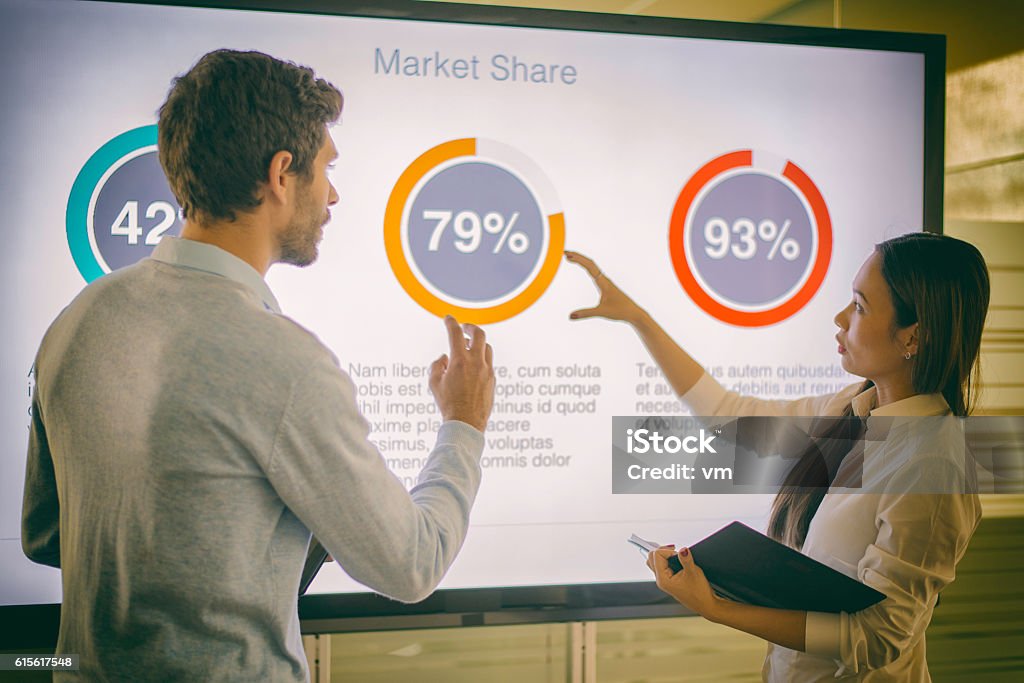 Businesswoman and businessman talking about market share Two business people standing in front of a presentation screen and talking about the graphs on it. They are showing market share percentages. Presentation - Speech Stock Photo