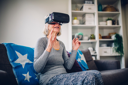 Senior woman sits on a sofa at home on her own. She is wearing a virtual reality headset with her hands in front of her