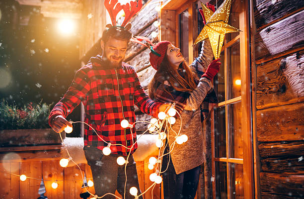 It's most wonderful time of the year Couple on vacation at mountain cabin. Decorating porch with string lights for Christmas. Wearing knitted sweaters, hats and scarfs. Austrian Alps. Evening or night with beautiful yellow lights lightning the scenes. decorating stock pictures, royalty-free photos & images