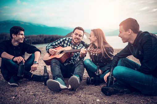 Four Smiling People with Acoustic Guitar Singing on Beach Camping. They are having summer fun on the sunrise.