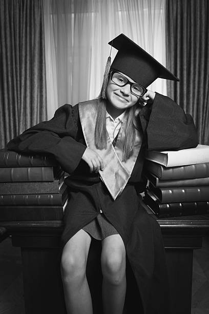 istock Smart girl in graduation cap posing on table at library 615638682