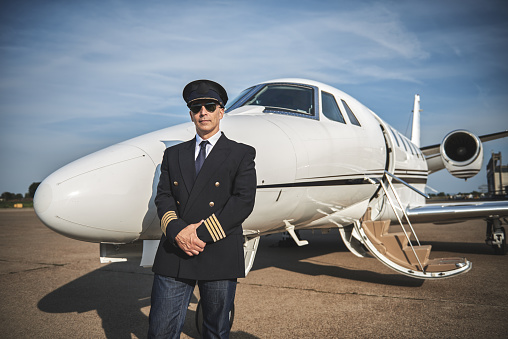 Portrait of a professional pilot in uniform and sunglasses standing next to the private jet airplane.
