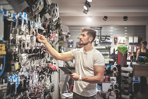 Young man is standing inside the bicycle store with digital tablet and looking at spare parts and equipment.