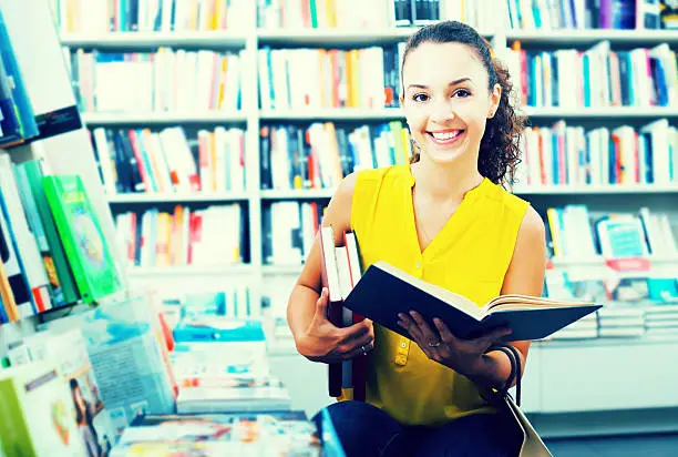 Portrait of positive young woman looking interested and reading textbook in book store