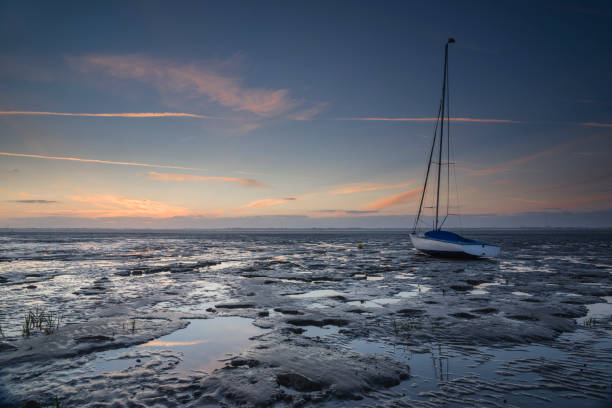 Sailboat in the wadden sea at dusk. Reflections of light summer clouds and blue sky in puddles. Taken in Dangast, Jade Bay, German North Sea Region, Friesland, Lower Saxony, Germany, Europe.