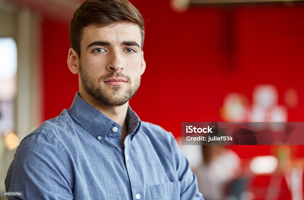 Confident male designer working in red creative office space Casual portrait of a business man showing determination and pride in a bright and sunny startup with the team in the background Serious Stock Photo
