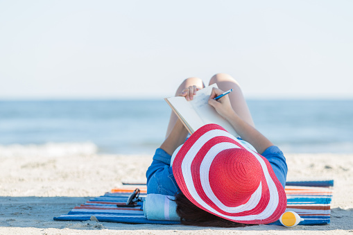 Peaceful woman writes in her journal while lying on the beach during a beach vacation. she is wearing a red and white striped straw hat. The ocean is blurred in the background.