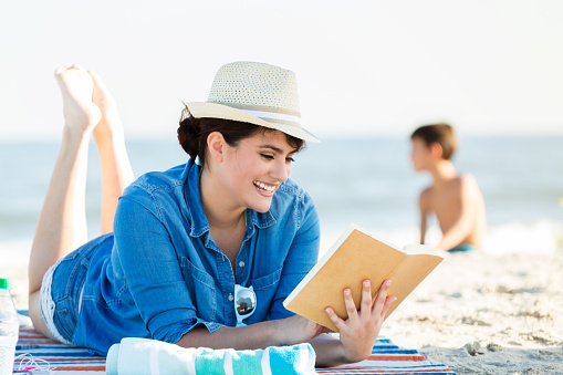 Beautiful content woman relaxes while reading book on the beach. She is wearing a denim shirt over her bathing suit. She is also wearing a straw hat. Her son is building a sand castle in the background.