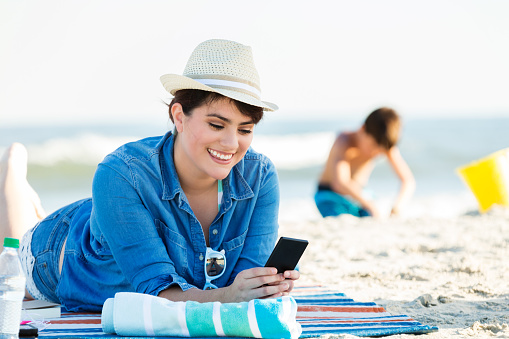 Hispanic woman smiles while video chatting or texting someone on the beach. She is lying on a beach towel or mat. She is wearing denim shirt over swimsuit. She is also wearing a straw hat. Her son is playing in the sand in the background.