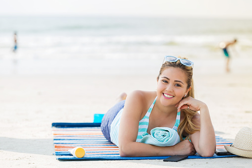 Pretty Hispanic woman enjoys sunbathing on the beach. She is laying on a beach towel or mat. A tube of sunscreen is beside her. People are playing in the background.