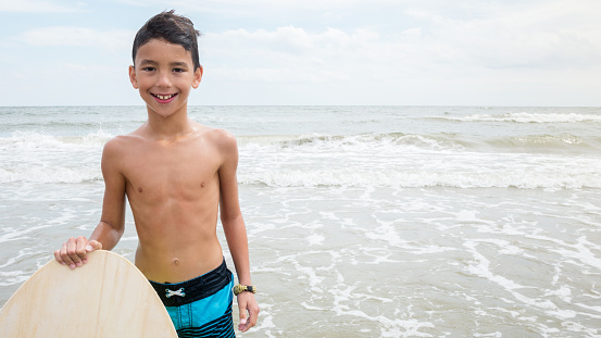 Happy Hispanic boy smiles cheerfully while holding a body board in the ocean. He has brown hair and is wearing a swimsuit. The waves are crashing in the background.