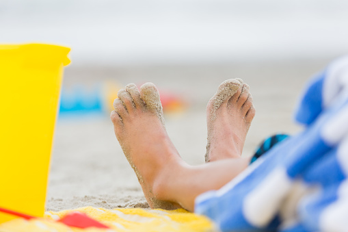 Close up of a boy's sandy feet at the beach. He is sitting in the sand. Beach toys are near his feet. He is wrapped in a beach towel.