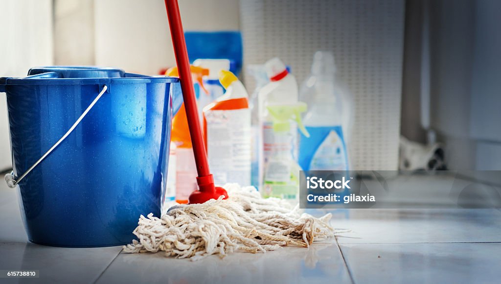 Cleaning products. Closeup of unrdcognizable home cleaning products with blue bucket and a mop in front in sharp focus. All products placed on white and poorly lit bathroom floor. Cleaning Stock Photo