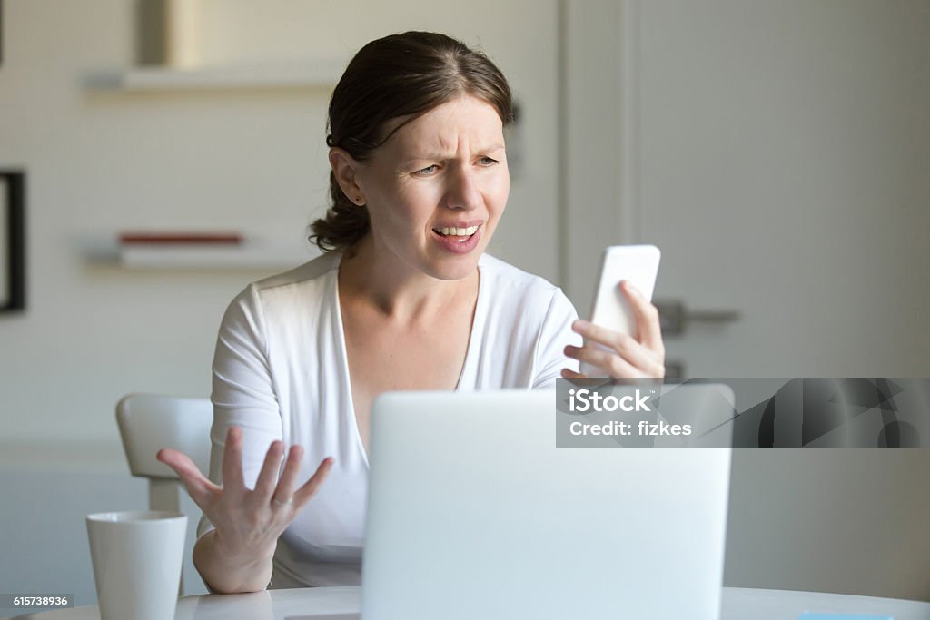 Portrait of a woman the laptop, looking screen of mobile Portrait of a young woman at the desk with a laptop, looking puzzled at the screen of the mobile she holds at her hand and message. Business concept photo, lifestyle Rudeness Stock Photo