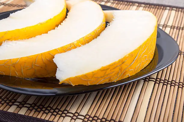 Three slices of juicy yellow melon on a black plate closeup on wooden background