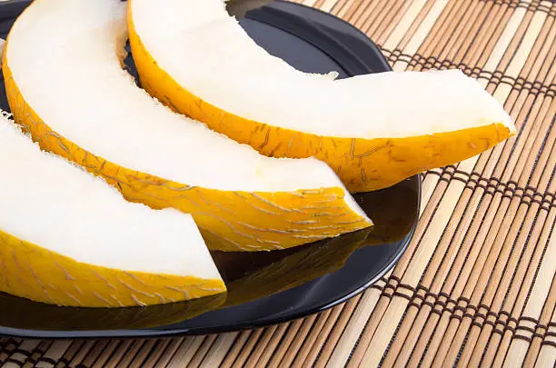View close-up on yellow juicy melon slices on a black plate on a striped wooden background