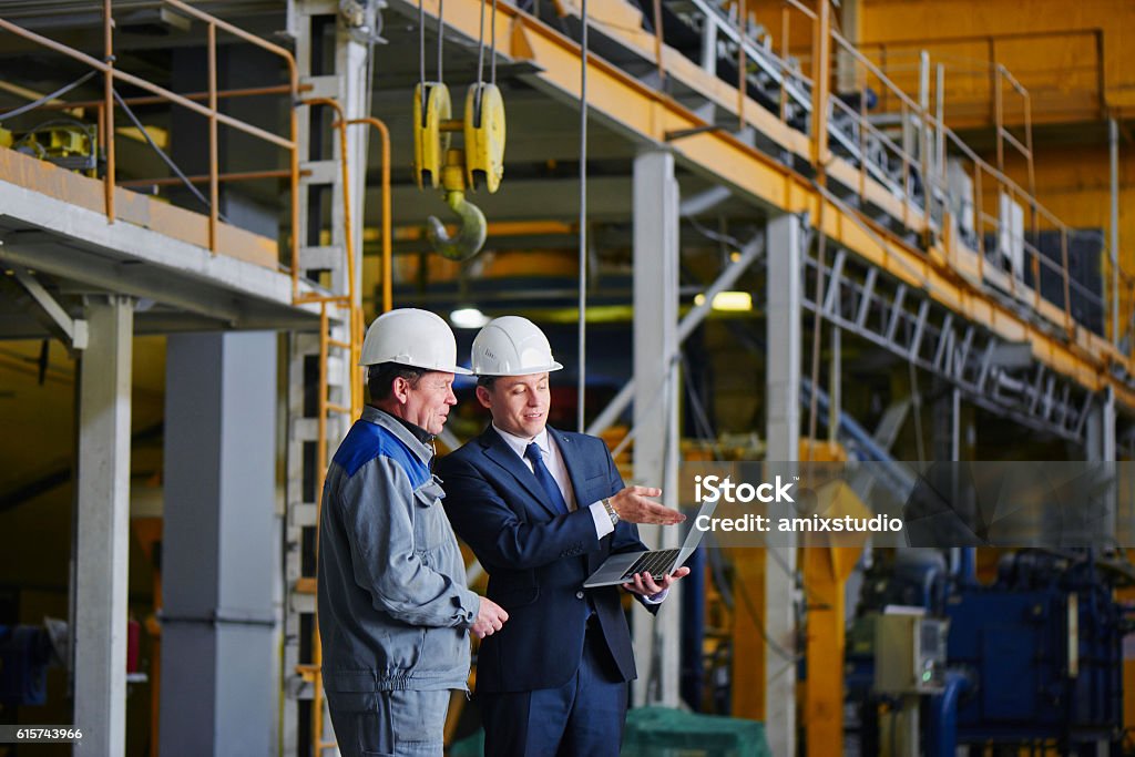 Man in a suit and a worker in overalls The man in the suit and the helmet holds the portable computer and shows up on the screen to the worker in overalls in an industrial building Office Stock Photo