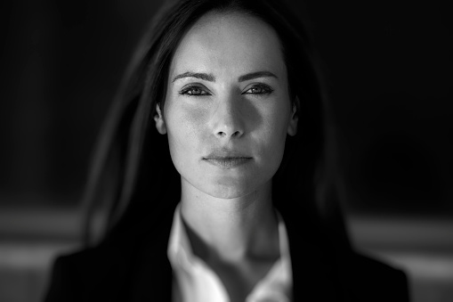 Portrait of a young woman with a pistol. She is sitting at a table. Her face is in shadow. A pistol lies in front of her in the faint light of an invisible ceiling light. It is the moment of deciding what to do next. Dark and mysterious.