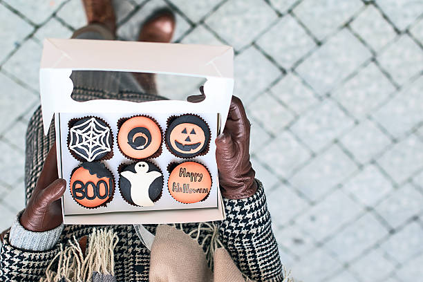 Woman holding a Halloween cupcakes box Young woman holding an open box full of Halloween styled cupcakes - cobweb, witch hat, carved pumpkin, boo, ghost and greeting black hat upside down stock pictures, royalty-free photos & images