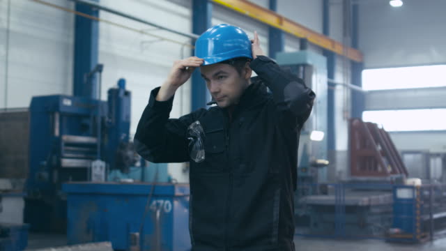 Factory worker in blue uniform is putting his hard hat and goggles on while walking.