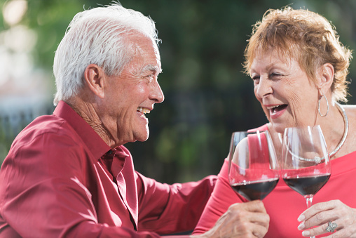 A loving senior couple sitting together at a table outdoors, face to face, smiling at each other, drinking red wine, laughing. The main focus is on the man.