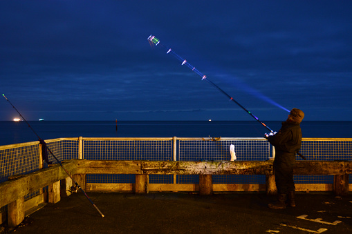 Shanklin, Isle of Wight, United Kingdom - December 6, 2014: The car park at Shanklin Beach makes a good spot for night fishing all year round. A man watches the end of his rod with a torch, hoping for a bite. Ships lights can be seen on the horizon.