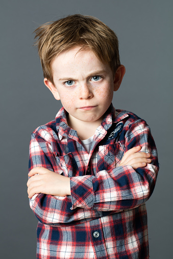 conflicted young 6-year old kid with messy red hair standing, crossing his arms to express his disagreement and frustration, grey background